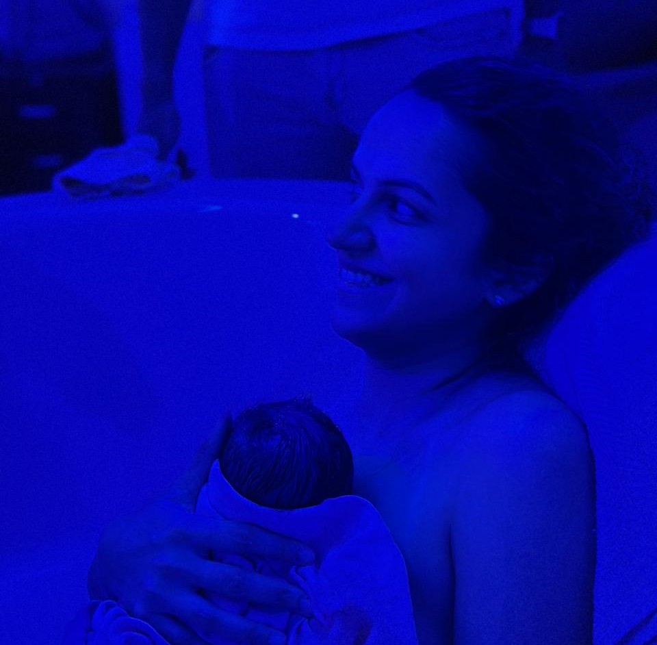 Hamsini smiling as she cradles her newborn in the dim blue light of the birthing room