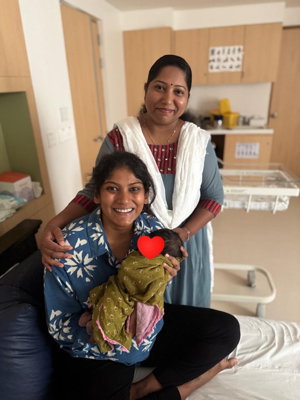 Vinita holding her newborn with a woman standing beside her in hospital, with the baby’s face covered by a heart