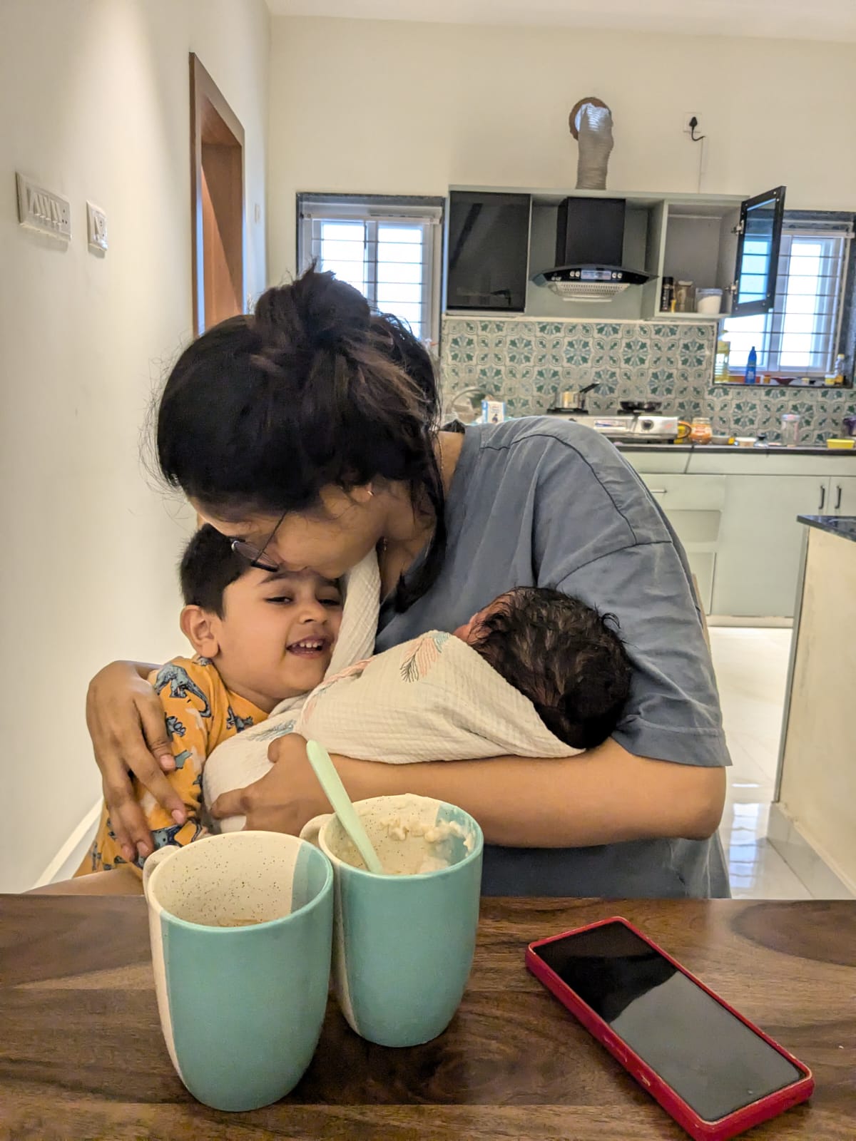Meera holding her newborn while hugging her older child at the kitchen table