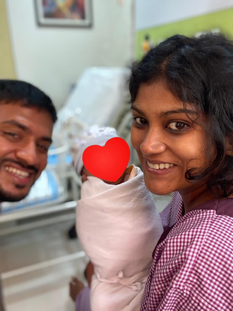 Vinita holding her newborn beside her smiling husband in hospital, with the baby’s face covered by a heart